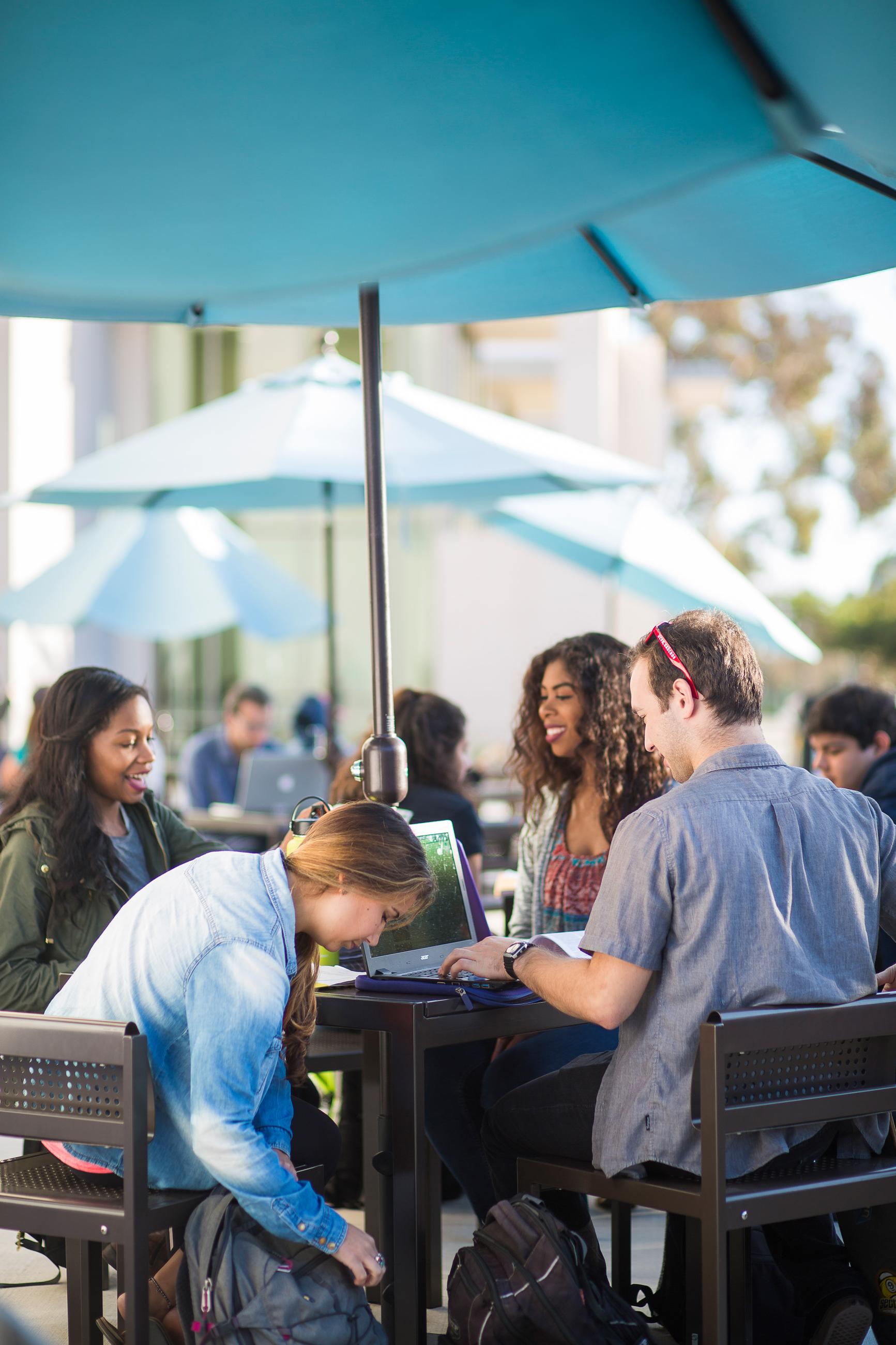 Students sitting around a table in an outdor seating area shielded from the sun with a large umbrella